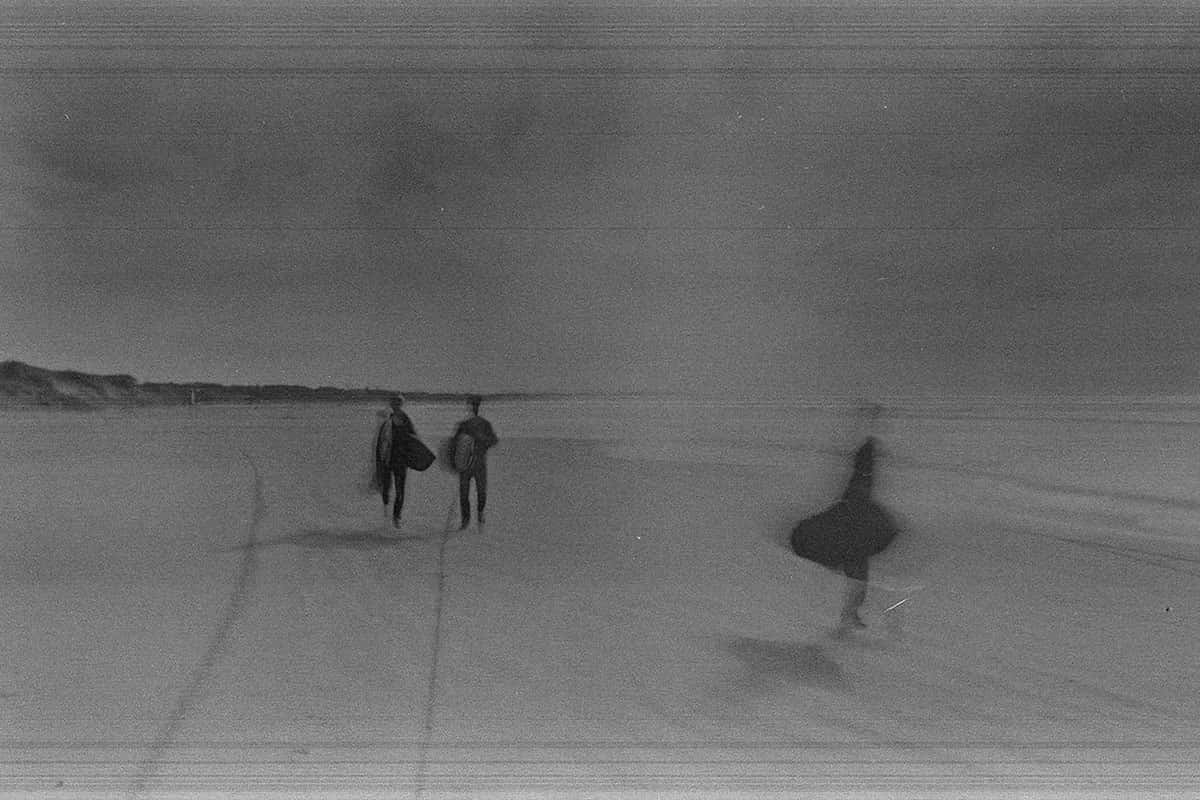 Fotografía en film 35 mm con grano marcado: tres surfistas caminan por la playa hacia el mar, siluetas difusas en luz plateada