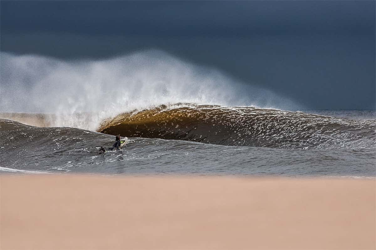Ola ámbar rompiendo en tubo bajo un cielo de tormenta gris oscuro, un surfista rema en la base y arena en primer plano