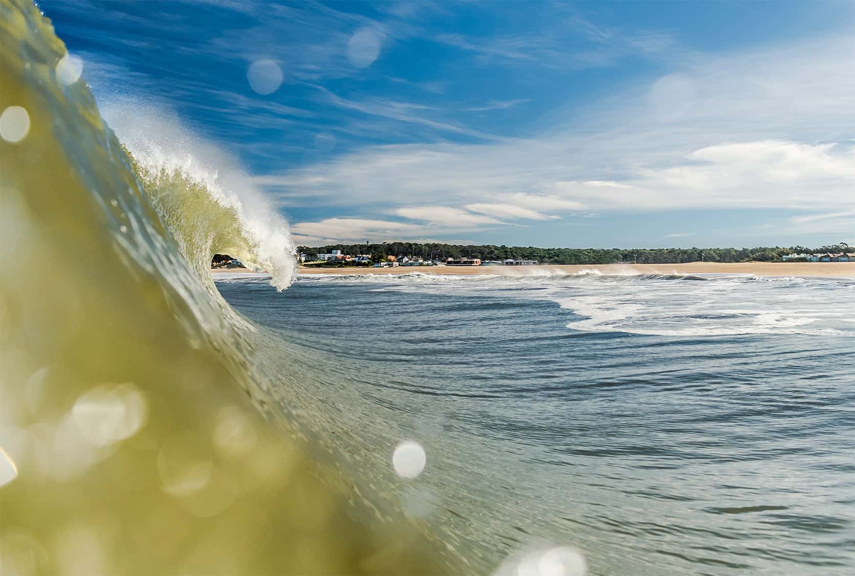 Ola verde rompiendo en primer plano fotografiada desde el agua, con playa y balneario uruguayo al fondo bajo cielo azul
