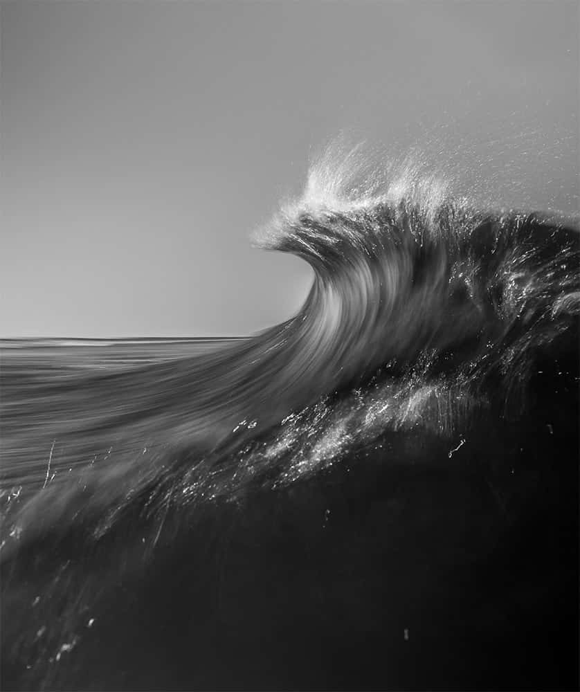 Fotografía vertical en blanco y negro de una cresta de ola en larga exposición, el agua barrida como humo contra cielo gris