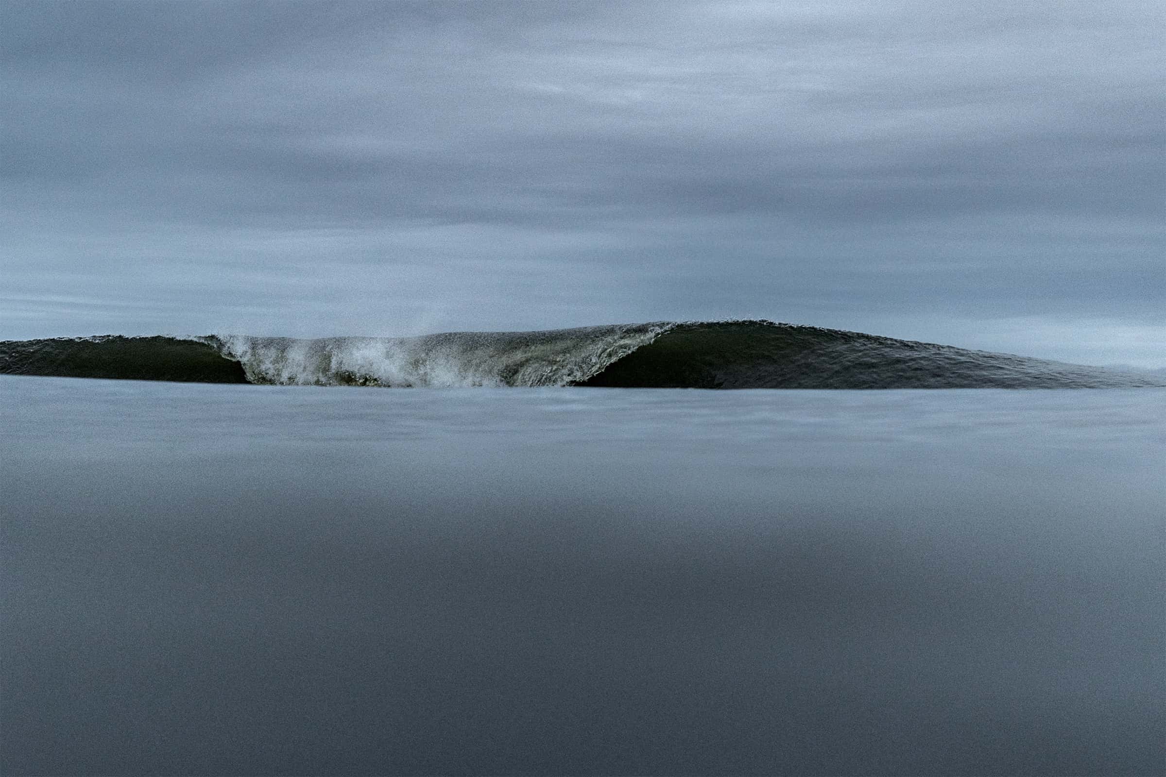 Ola verde rompiendo en primer plano fotografiada desde el agua, con playa y balneario uruguayo al fondo bajo cielo azul