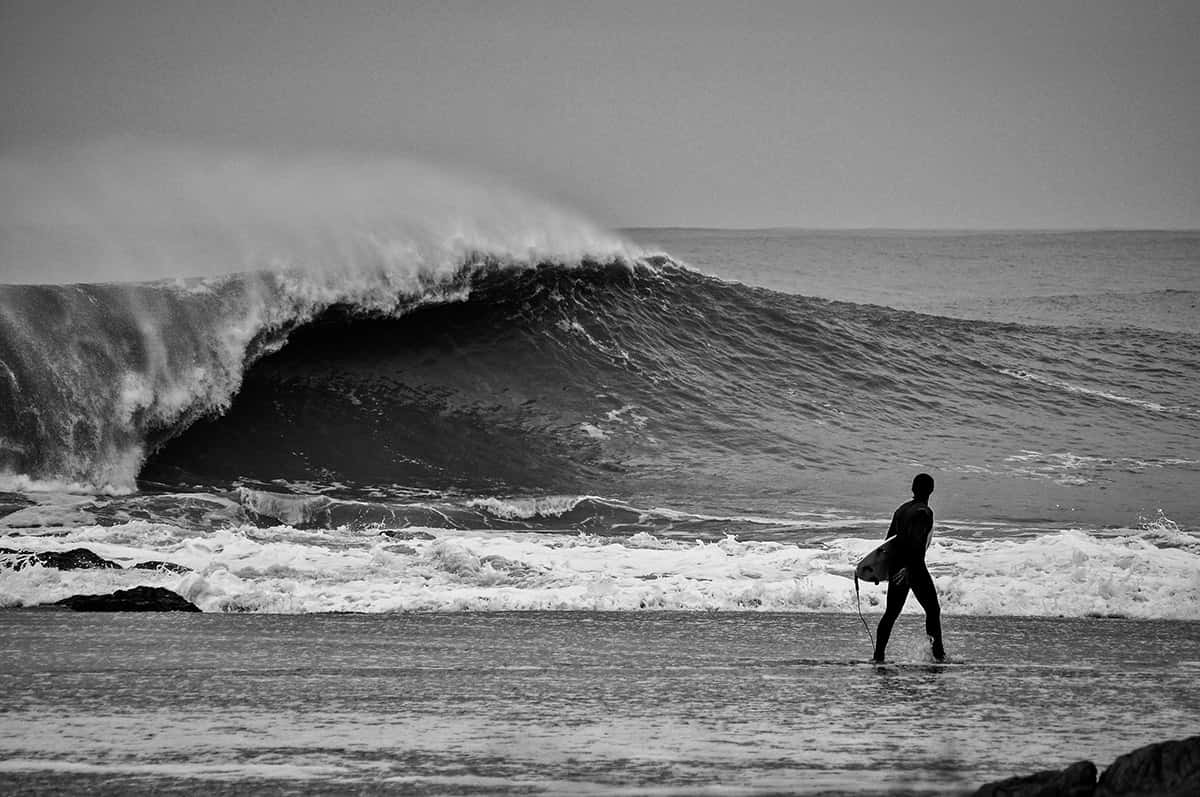 Fotografía en blanco y negro: un surfista camina por la orilla con la tabla bajo el brazo mientras una ola grande rompe detrás